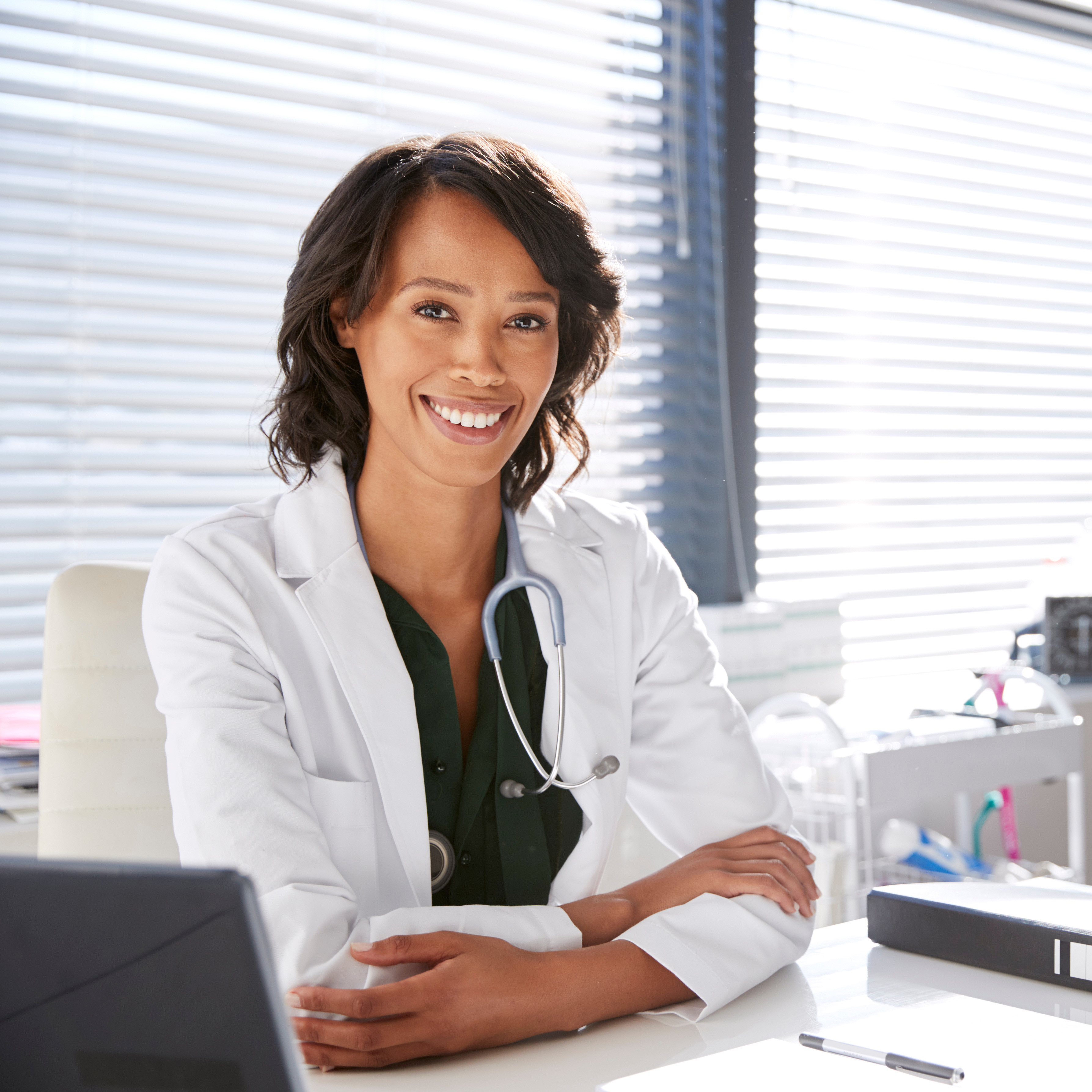 Portrait Of Smiling Female Doctor Wearing White Coat With Stethoscope Sitting Behind Desk In Office.